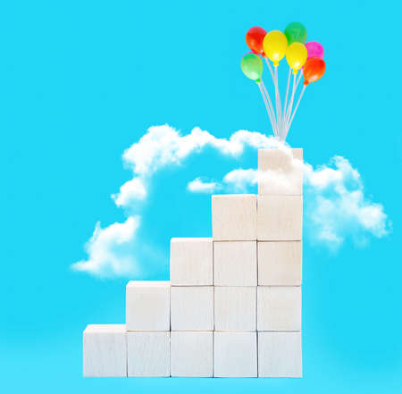 Wooden toy blocks stacked into stairs leading to the clouds with colorful party balloons on top against blue background. The concept of taking stairs to success.の写真素材