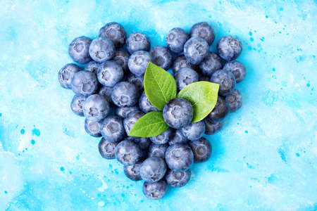 Heart symbol made from fresh ripe blueberries and green leaves on a light blue watercolor background. Healthy eating and immunity boost concept.の写真素材