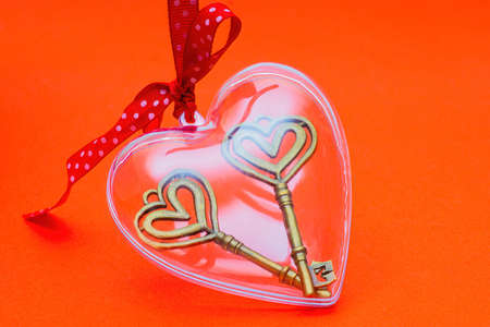 Two retro style skeleton keys in a clear heart shaped box with a red dotted ribbon bow on a red background. The concept of openness and clarity for a solid relationship.の写真素材