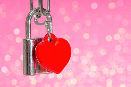 Close-up of a locked padlock with a red heart shaped pendant against a pink background. Romantic St....