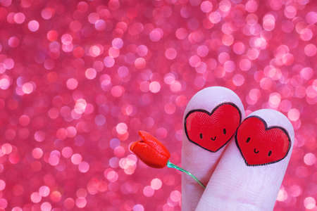 Crossed fingers with smiling heart-shaped faces holding a miniature toy rose against a bokeh...
