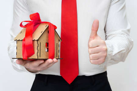 Businessman holding a miniature house model wrapped with a red ribbon giving a thumbs-up. Home loan approval concept.の写真素材