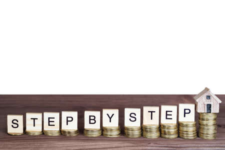 Wording STEP BY STEP made from wooden letter blocks and a miniature house placed on ascending coins...