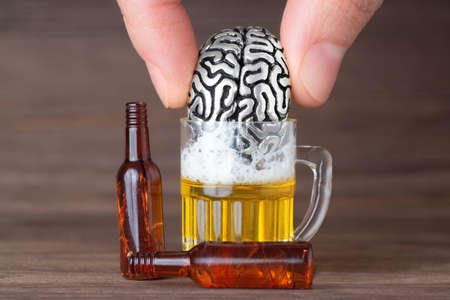 Fingers drowning a miniature anatomical steel copy of human brain in a small beer mug against a wooden background. Loss and grief leading to substance abuse.の写真素材