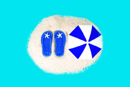 Miniature lip flops and sun umbrella on a pile of sand isolated on a blue background. Creative sandy...