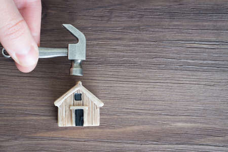 Fingers holding a miniature steel hammer over a tiny toy house model against a wooden background. Home renovation project.の写真素材
