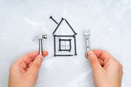 Hands holding a miniature steel hammer and a wrench by a house shape made from black colored screws on a concrete background. Creative home repair service concept.の写真素材