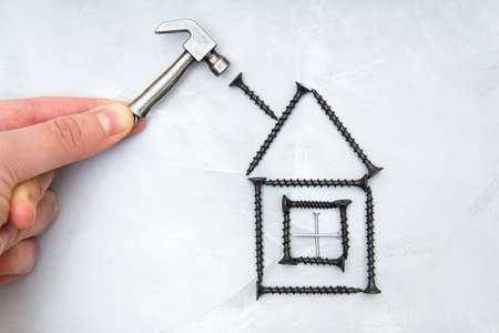 Hand holding a miniature steel hammer by a house shape made from black colored screws on a concrete background. Home repair and improvement concept.の写真素材