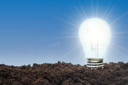 Glowing incandescent light bulb sticking out of the soil against a blue sky background. Green energy...