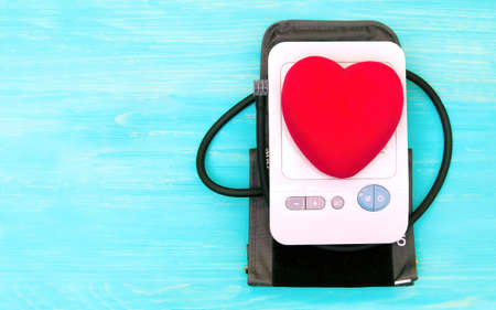 Large red heart shape on top of a blood pressure monitor placed on a turquoise background with copy...