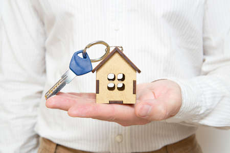 Man holding a house key with a miniature wooden house model keychain offering it to the viewer. Real...