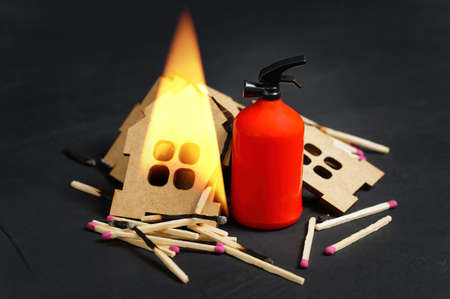Miniature fire extinguisher and matchsticks placed by a collapsed burning toy house on black background.の写真素材