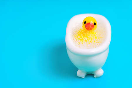 Tiny yellow duck in a toy bathtub full of soap bubbles isolated on blue background with copy space. Hygiene habits for kids.の写真素材