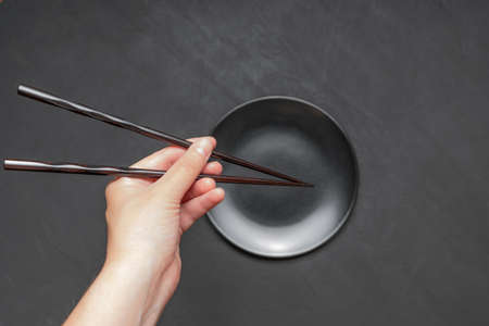 Pair of lacquered chopsticks in hand over an empty matte black plate on dark background.の写真素材