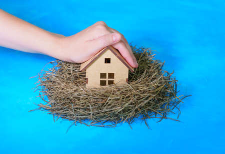 Hand covers a miniature wooden house placed in a hay nest isolated on blue background. Home insurance concept.の写真素材