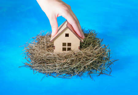 Hand placing a miniature wooden house in a hay nest isolated on blue background. Creative family house concept.の写真素材