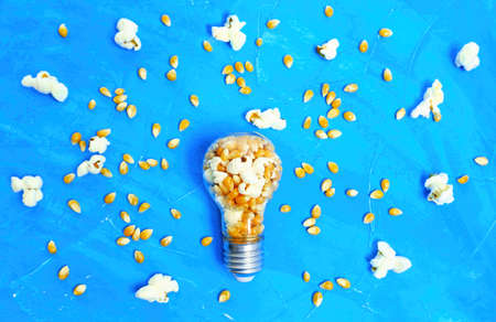 Lightbulb full of raw and popped corn kernels on blue background. Creative seed funding return...