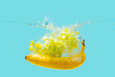 Bunch of white grapes and yellow banana dropped in water with splashes isolated on blue background.の写真素材
