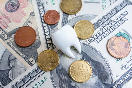 White tooth lies on a dollar bills and euro coins background. Dental insurance related concept.