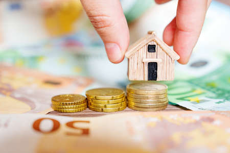 Hand tanking a small toy house model away from a stack of coins framed with euro banknotes, selective focus. Creative foreclosure concept.の写真素材