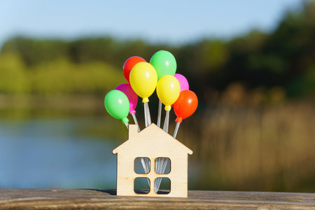 Miniature flat house shape with a bunch of small colorful balloons against a beautiful lake landscape background. Creative moving in party concept.の写真素材