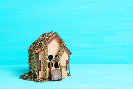 Toy timber house wrapped with bronze-toned chain and secured with a massive padlock isolated on blue background with copy space.の写真素材