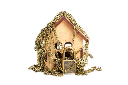 Wooden toy house wrapped in chains with a bronze-toned padlock isolated on white.の写真素材