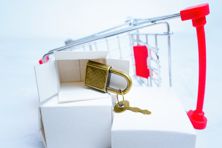 Small bronze toned padlock with key falling of a white delivery box placed by an overturned miniature push trolley. Security hardware store concept.の写真素材