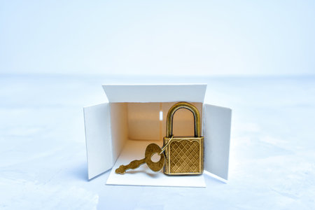 Close-up of a bronze tone padlock and key by an empty white box on a neutral background. Security...