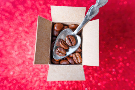 Taking roasted coffee beans with a small teaspoon out of a miniature shipping box isolated on red...