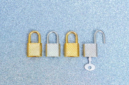 Row of four silver and golden toned padlocks on a glittering background. One padlock is opened with a key.の写真素材