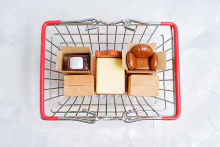 Selection of essential home furniture figurines and tiny delivery boxes placed in a shopping cart, top view. New apartment furnishing concept.の写真素材