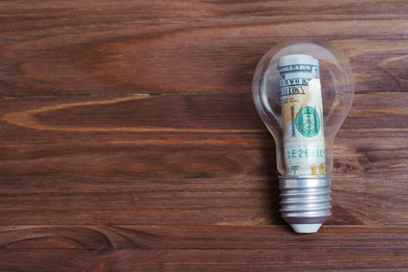 Hundred dollar bill rolled inside a light bulb isolated on a wooden table with copy space. Creative...