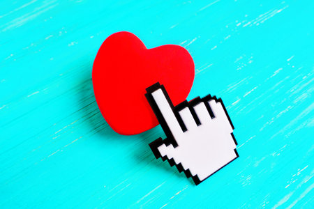 Large pixelated hand-shaped cursor touches a red heart placed on a light blue wooden background....
