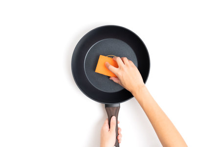 Crop view of a pair of female hands holding a frying pan and cleaning it using an orange sponge isolated on white. Domestic chores, household tasks, kitchen cleaning and general housework concept.の写真素材