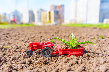 Farming vs Urbanization: Red toy tractor with a trailer filled with young green shoots standing on freshly plowed land, with a blurred image of a cityscape in the background.の写真素材
