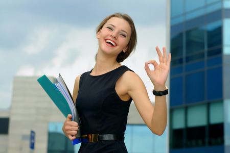 Business woman with a folder in her hand shows that everything is OK and fun laughs against a background of modern building facadesの写真素材