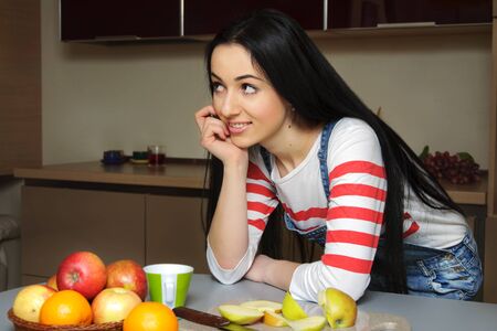Brunette housewife wearing in blue denim overalls enthusiastically listens to someone. Girl looking slightly to the side and rests his hand on the table. の写真素材