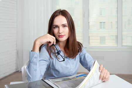 The girl sits at a table in an office holding a newspaper in one hand and in the other glassesの写真素材
