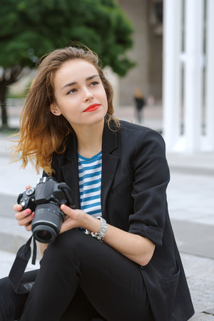 Woman photographer sits on the steps with a camera in her hands and looks asideの写真素材