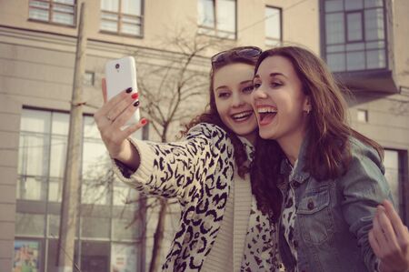 Photo in vintage style. Two girls taking pictures of themselves and laughing at city background.の写真素材
