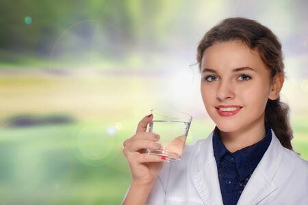 Woman doctor in a white coat smiling and drinking water from a glass cup against a blurred background in green and yellow colorsの写真素材