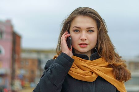 Girl in a black coat and orange scarf talking on phone on the background of the cityscapeの写真素材
