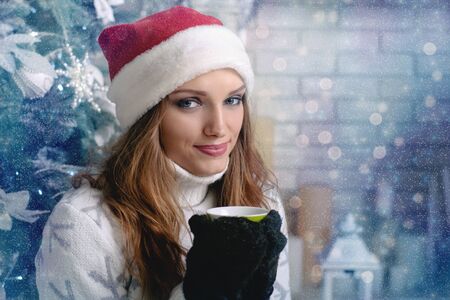 Woman in a warm sweater, hat and mittens drinking a hot drink against a blurred background Christmas treeの写真素材