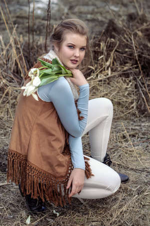 Girl in fashionable clothes sitting against a background of dry branches and holding a wilted bouquetの写真素材