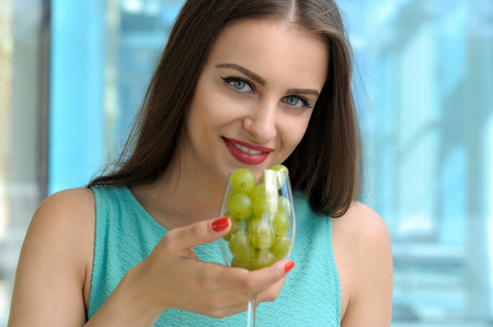 Woman brings to her mouth a wine glass full of ripe green grapes. She has long dark hair, clear skin and dressed in a turquoise dress.の写真素材