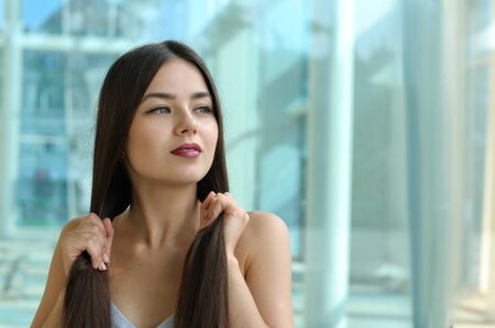 Beautiful girl touches her hair. She has long brown hair and well-groomed skin. Portrait against glass and metal.の写真素材