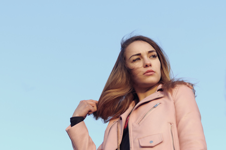 Fashion portrait of a beautiful girl against a blue sky. She is dressed in a leather pink jacket and she straightens her hair with her hand.の写真素材