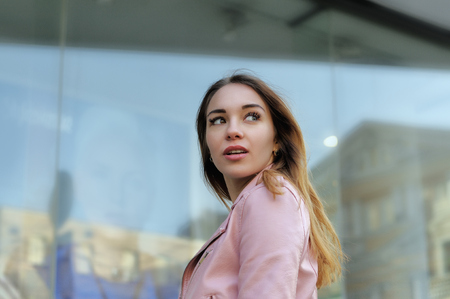 Beautiful girl turned on the background of the glass shop windows. She is dressed in a pink leather jacket and she has long beautiful hair.の写真素材