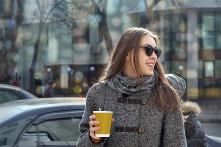 Business lady goes to work with a cup of coffee early in the morning on a busy street. She is dressed in a gray coat and gray scarf, she is in a good mood and she smiles, looking backの写真素材